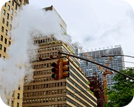 Street intersection with traffic light and steam rising from a vent, surrounded by tall city buildings.