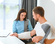 Woman supporting man with an arm cast using crutches, sitting together indoors by a window.