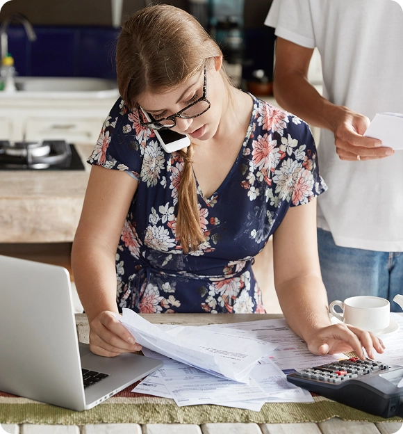 Woman in a floral dress talking on the phone while reviewing bills and using a calculator at a kitchen table with a laptop and a man holding papers nearby.