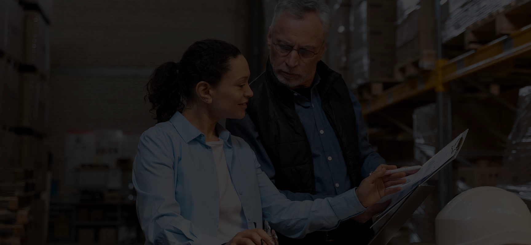 Two warehouse workers reviewing documents on a clipboard among stacked pallets and shelves.