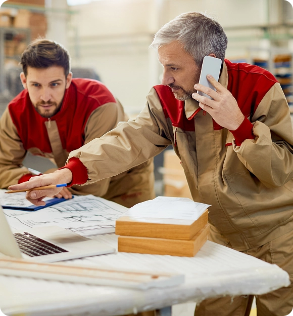 Two men in work uniforms reviewing architectural plans and pointing at a laptop, one speaking on a smartphone.