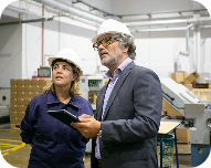 Two factory workers wearing white helmets discussing work inside a warehouse, one holding a digital tablet.
