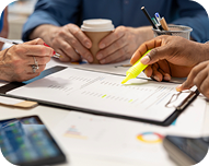 Three people reviewing printed documents with one person highlighting text in yellow and another holding a coffee cup.