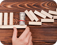 Hand stopping a row of falling dominoes with a wooden block wrapped with a hundred-dollar bill.