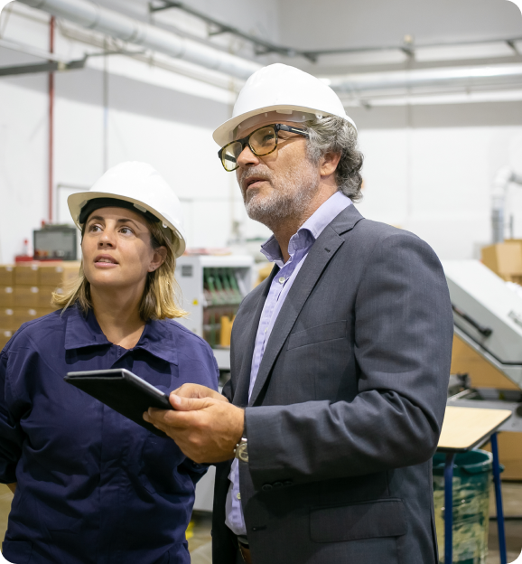 Two professionals in a warehouse wearing white hard hats, one holding a tablet and both looking intently at something off-frame.