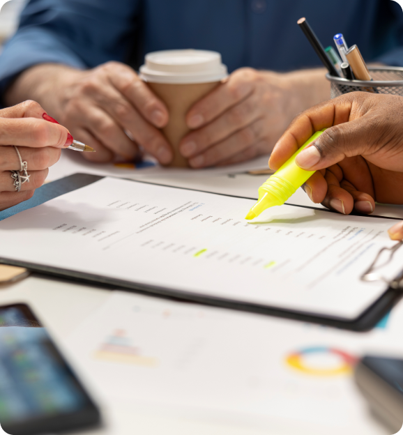 Hands of people working on documents with one person highlighting text with a yellow marker and another holding a pen and coffee cup.