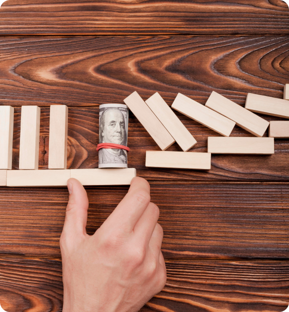 Hand placing a wooden block to stop falling dominoes, with a rolled hundred-dollar bill held by a rubber band acting as a barrier.