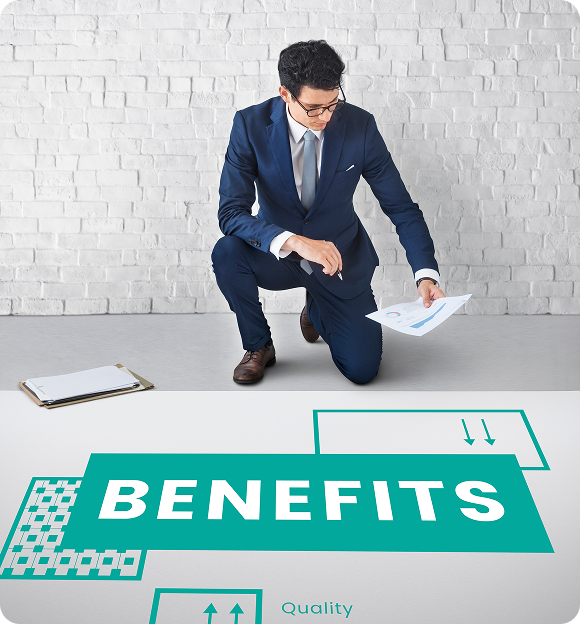 Man in blue suit kneeling on one knee, examining documents near a large graphic with the word 'BENEFITS' on a white brick background.