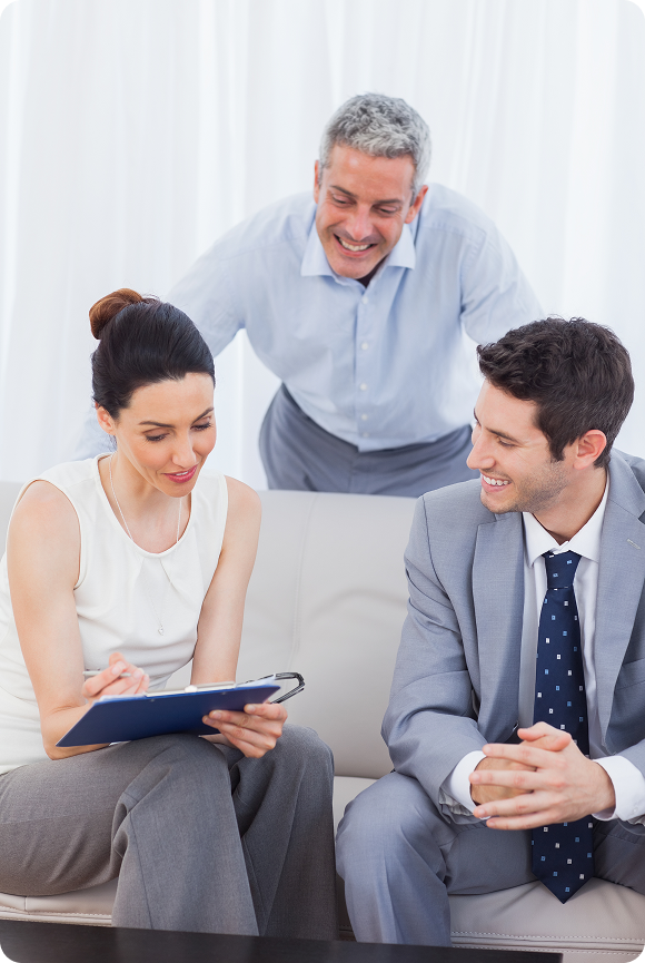 Three business professionals seated on a sofa reviewing a document on a clipboard, smiling and engaged in discussion.