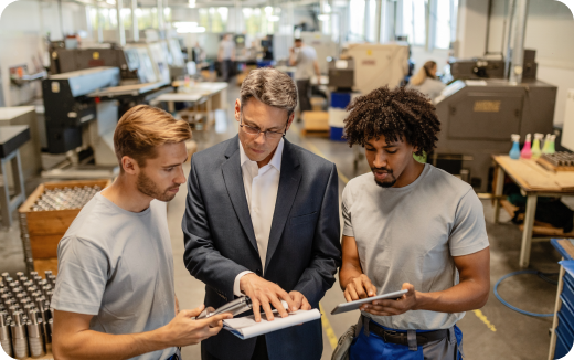 A man in a suit discusses a document with two workers holding tablets inside a manufacturing facility.