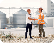 Two construction workers wearing hard hats and safety vests shaking hands at an industrial site with storage tanks in the background.