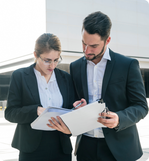 Two business professionals in suits reviewing documents on a clipboard outdoors.