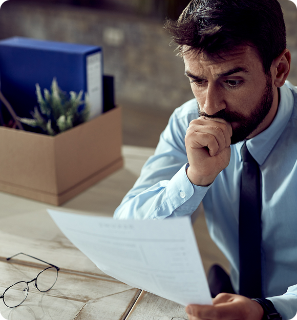 Worried man in a dress shirt and tie reading a document at a desk with a box of personal items and glasses nearby.