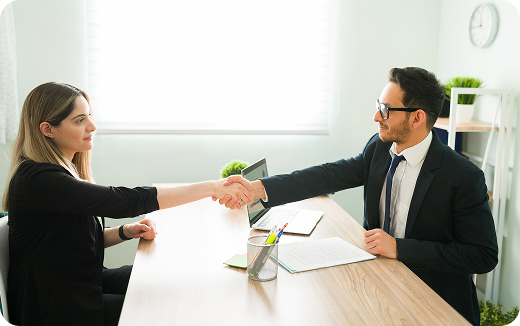 A man and woman in business attire shaking hands across a desk in a bright office setting.