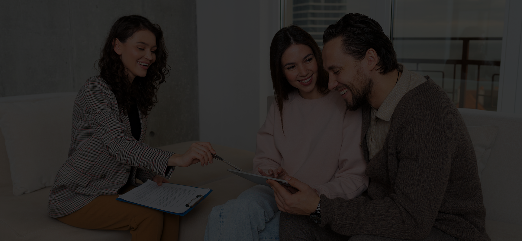 A professional woman with a clipboard explains documents to a smiling couple reviewing a tablet together on a couch.