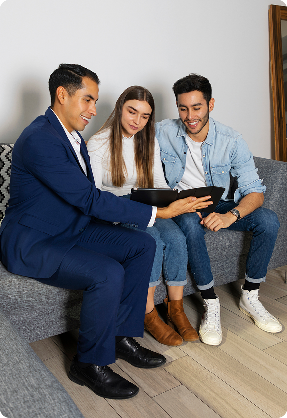 Three people sitting on a gray couch, looking at a tablet with interest and smiling.