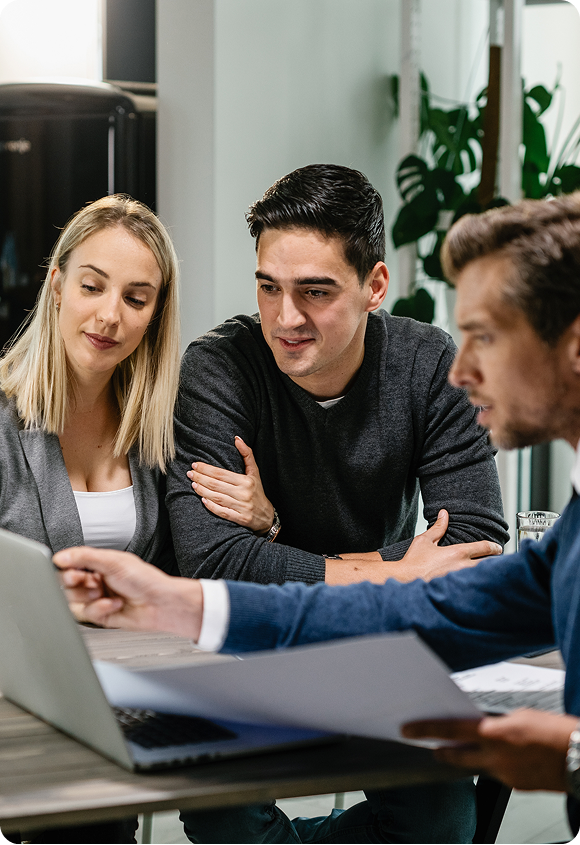 A man in a blue sweater explains something on a laptop to a couple seated at a table in a modern office.