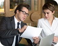 Man in glasses explaining document details to woman working on a laptop in an office setting.