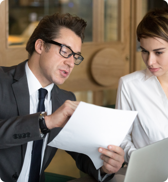 Man in suit and glasses discussing a document with a woman in a white shirt in an office setting.