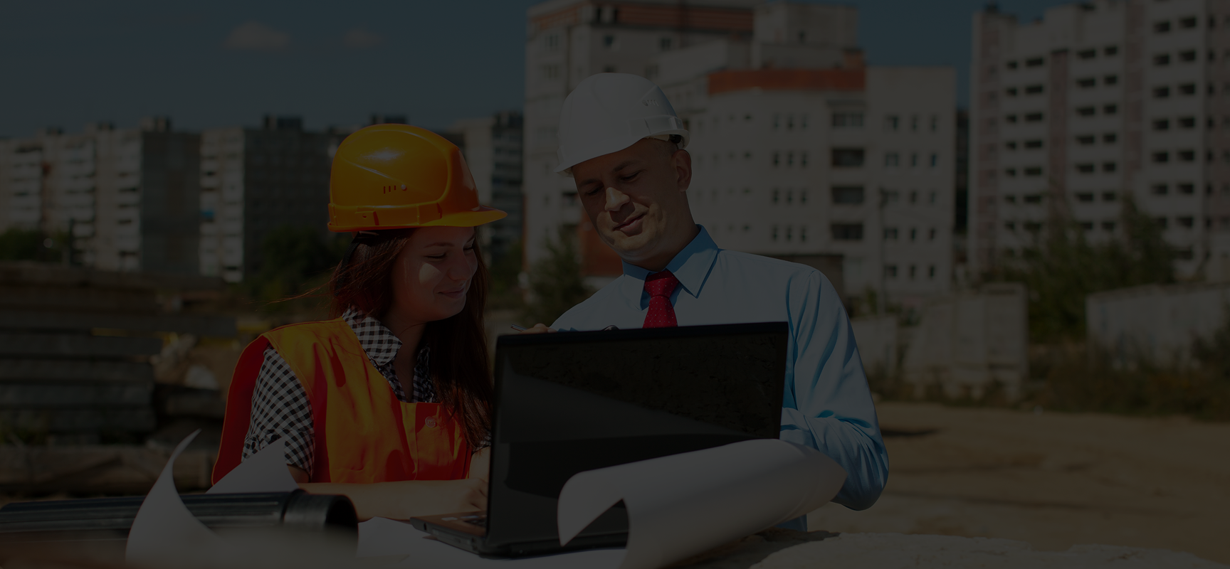 Two construction professionals reviewing plans on a laptop at a building site with urban apartment buildings in the background.