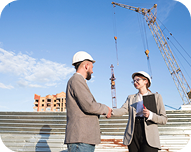 Two construction professionals wearing helmets shaking hands at a building site with cranes and partially built structures in the background.