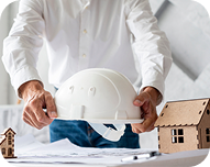 Person holding a white safety helmet above architectural blueprints on a table with wooden house models.