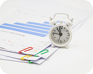 Small white alarm clock showing 9:12 placed on printed charts with colored paper clips.