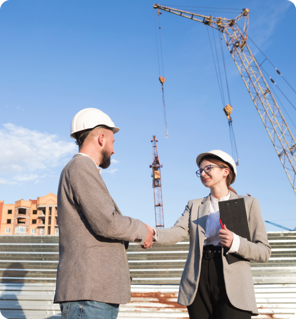 Two construction professionals wearing hard hats shaking hands at a building site with cranes and a partially built structure in the background.