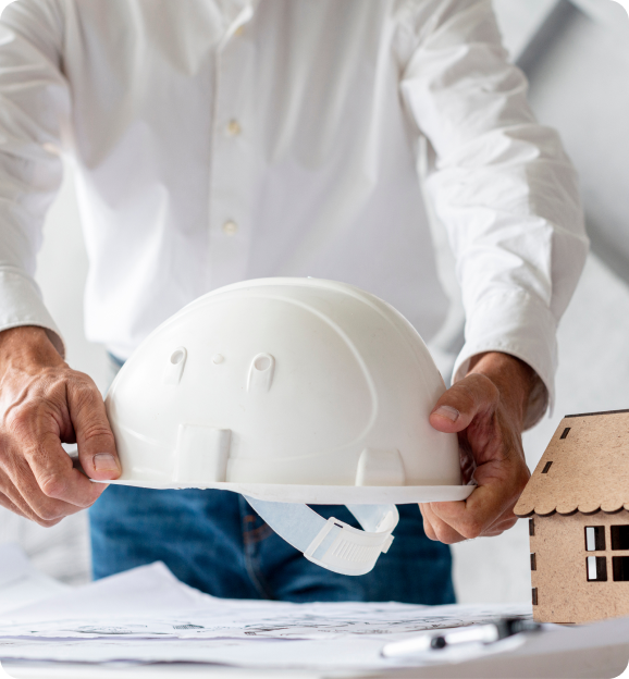 Person in white shirt holding a white construction hard hat over architectural blueprints with a small wooden house model on the table.