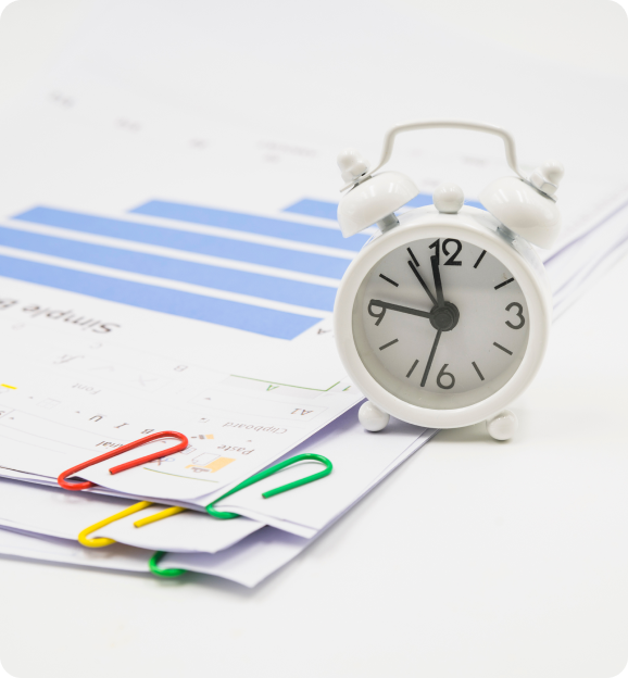 White alarm clock showing 11:57 next to documents clipped with red, green, and yellow paperclips on a white surface.