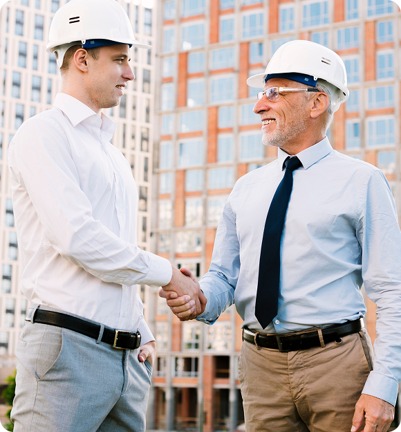 Two construction professionals wearing white hard hats shaking hands in front of a building.