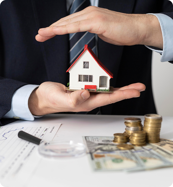 Person in suit holding a small house model between their hands with stacks of coins and dollar bills on a desk.