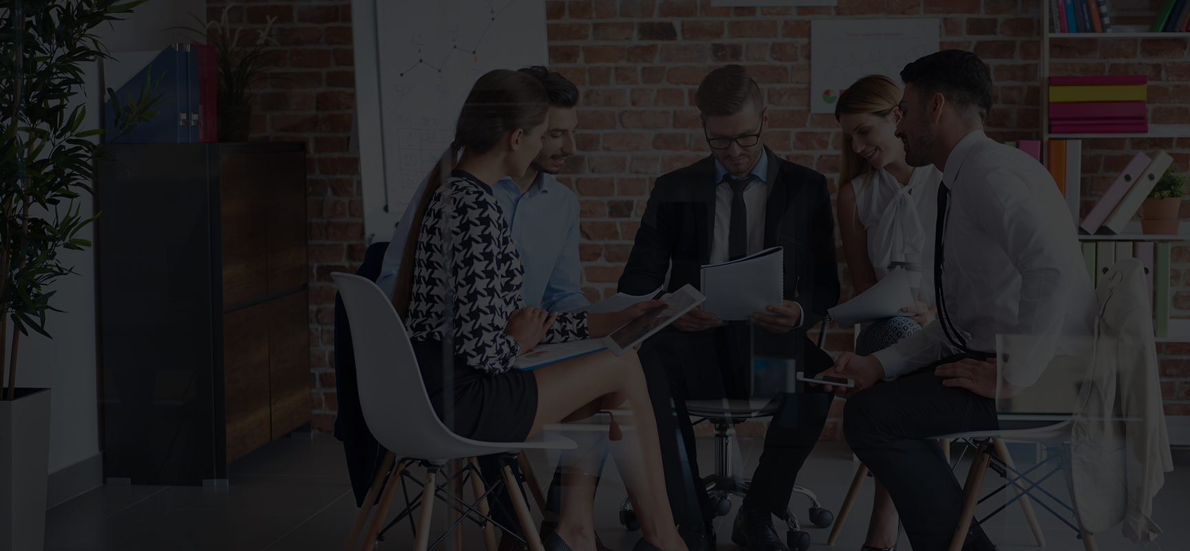 Five professionals in business attire seated in a modern office, discussing documents in a collaborative meeting.