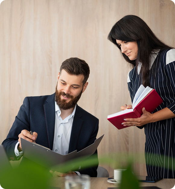 Man in a suit signing a document while a woman holding a red notebook looks on.