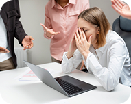 Stressed woman covering her face sitting at a desk with a laptop while colleagues gesture around her.