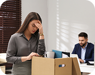 Stressed woman packing her belongings in a box with a man working on a laptop in the background.