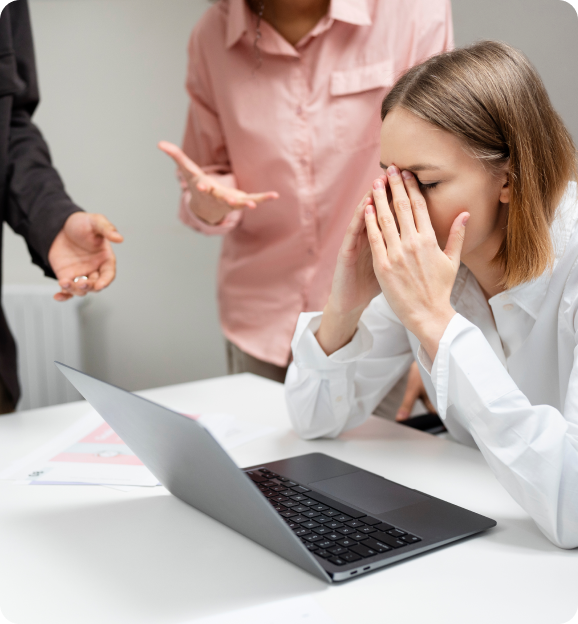 Stressed woman sitting at a desk with a laptop, covering her face with her hands, while two people in the background gesture expressing frustration.