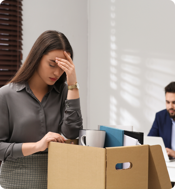 Stressed woman holding her forehead standing by a box of office supplies indicating possible job loss.