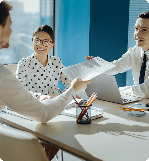Two men and a woman in a business meeting exchanging a document across a table with a laptop and office supplies.