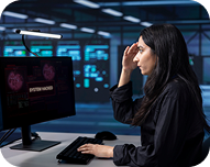 Woman in dark clothing looking stressed while working on a computer with cybersecurity data displayed on the screen in a dimly lit control room.
