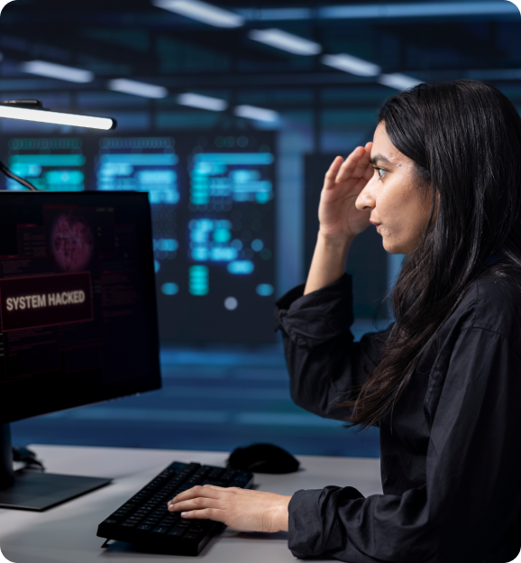 Woman with long dark hair looking worriedly at a computer screen displaying a 'SYSTEM HACKED' warning in a dimly lit control room.