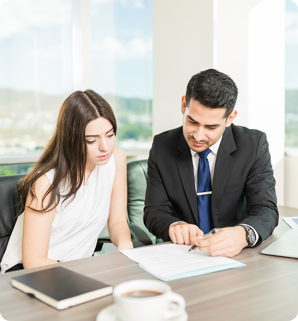 Man in suit explaining documents to a woman in a white sleeveless top at a desk with a notebook and coffee cup.