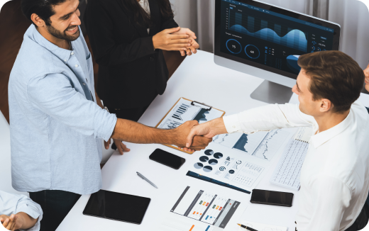 Two men in office shaking hands over a desk with charts, documents, tablets, and computer screen showing data graphs.