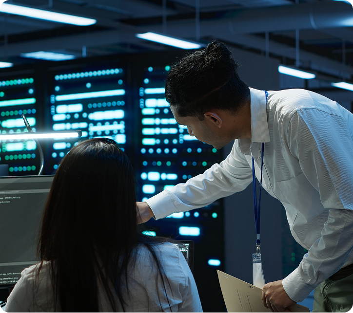 IT professionals collaborating in a server room, with one pointing at a computer screen displaying code.