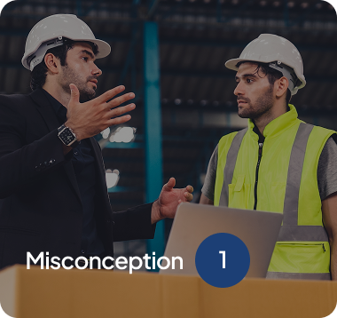 Two male workers wearing white safety helmets discussing in a warehouse, one in a black suit gesturing hands, the other in a yellow safety vest listening.