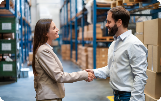 A man and a woman shaking hands and smiling in a warehouse aisle with shelves and boxes.