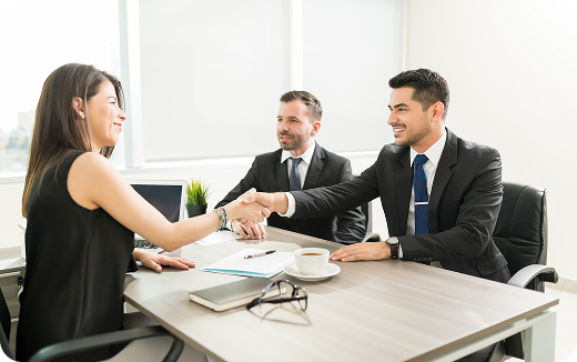 Two businessmen in suits shaking hands with a woman across a table in a bright office.
