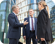 Three business professionals in suits having a discussion outside a modern glass office building.