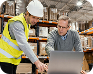 Two men, one in a safety vest and helmet, discussing inventory in a warehouse while looking at a laptop.