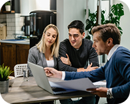 Three people sitting at a table in a modern office looking at a laptop screen together.